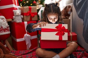 Obraz na płótnie Canvas Adorable hispanic girl unpacking gift sitting on floor by christmas tree at home