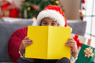 Adorable hispanic girl covering face with book sitting on sofa by christmas tree at home