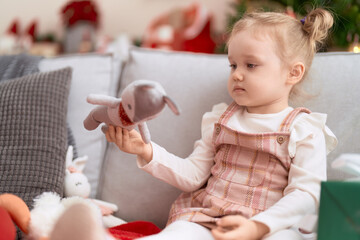 Fototapeta premium Adorable caucasian girl holding toy sitting on sofa by christmas tree at home