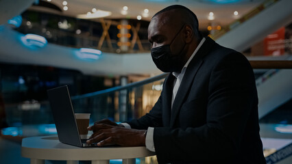 Middle-aged adult man worker manager boss company CEO businessman employer entrepreneur sitting in cafe with laptop working online project wearing medical face mask COVID-19 coronavirus pandemic work