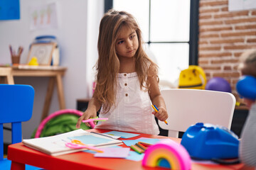 Adorable hispanic girl student sitting on table holding pencil color and scissors at kindergarten