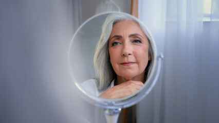 Mirror reflection female wrinkled face 50s middle-aged Caucasian woman senior lady looking self...