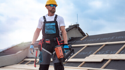 A carpenter with tool belt is holding a electric nailer  in his hand on-the-job on the roof.