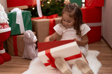 Obraz na płótnie Canvas Adorable hispanic girl unpacking gift sitting on floor by christmas tree at home