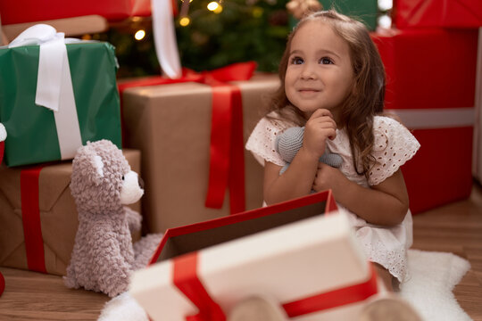 Adorable hispanic girl hugging teddy bear sitting on floor by christmas tree at home