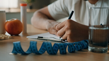 Close up male nutritionist unrecognizable man sport fitness trainer sitting at table writing prescription healthy nutrition fitness diet on paper count calories with apple vitamins pills tape measure