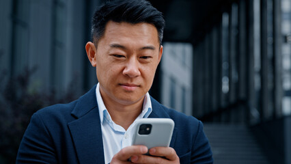 Close up Asian 40s businessman with smartphone texting browsing smiling looking at camera standing outdoors in city. Middle-aged man CEO has business chat using mobile phone check news in social net