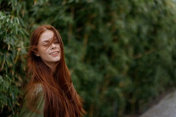 Portrait of a beautiful young girl in the city with a beautiful smile and red hair in a green raincoat in the city against a background of bamboo in spring, lifestyle in the city