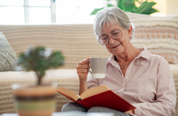 Handsome senior woman sitting on the floor at home reading a book while enjoying a cup of tea....