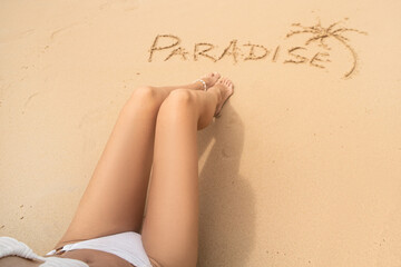 Word paradise and one palm on the sandy beach near the women's legs. Summer vacation concept. Realax.