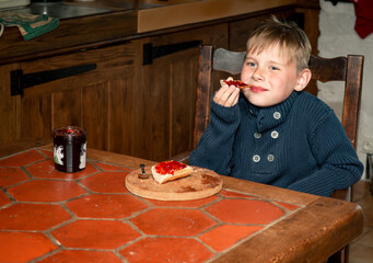 a boy of eight years old eats cherry jam with bread at the table in the kitchen
