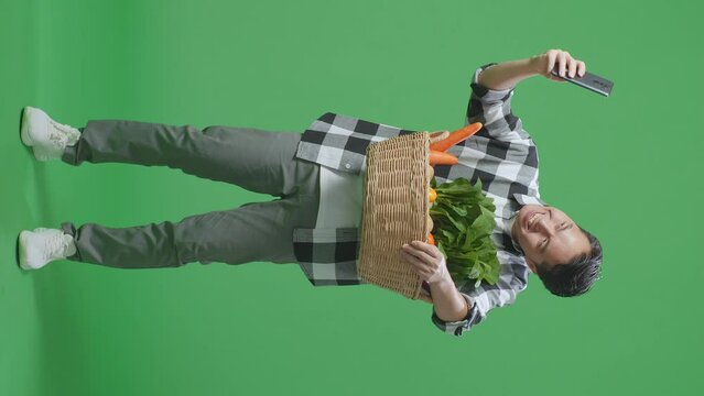 Full Body Of Asian Male Farmer With Vegetable Basket Taking Selfie By Smart Phone On Green Screen Background In The Studio
