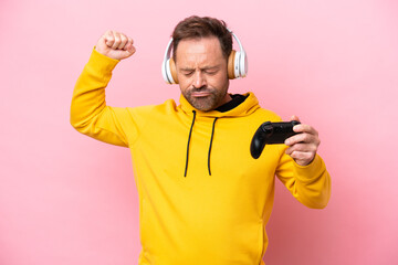 Middle age man playing with a video game controller isolated on pink background doing strong gesture