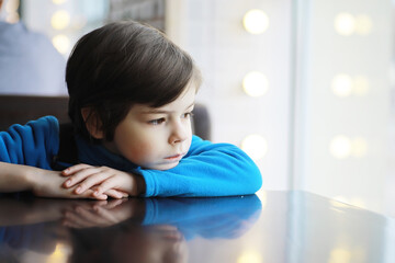 A young boy sits at a table near the window in a cafe and waits for a waiter to come up and take an...