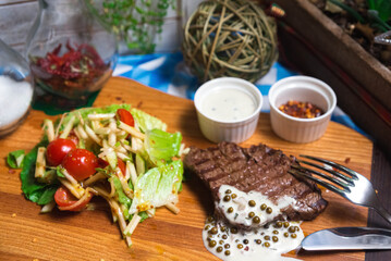 A piece of grilled beef with green peppercorn sauce on a wooden board with a salad