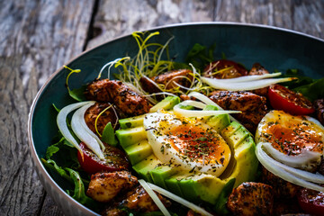 Tasty salad - fried chicken breast, avocado, boiled eggs, mini tomatoes and fresh green vegetables on wooden background
