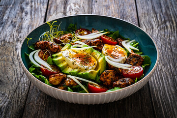 Tasty salad - fried chicken breast, avocado, boiled eggs, mini tomatoes and fresh green vegetables on wooden background

