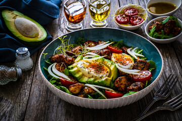 Tasty salad - fried chicken breast, avocado, boiled eggs, mini tomatoes and fresh green vegetables on wooden background
