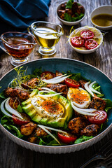 Tasty salad - fried chicken breast, avocado, boiled eggs, mini tomatoes and fresh green vegetables on wooden background
