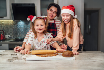 Laughing little girls in red Santa hat with mother family portrat. They making Christmas gingerbread cokies using rolling pin together in home kitchen. Happy holidays preparation and childhood concept