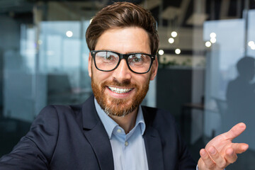 Businessman looks into smartphone camera and talks on video call, man uses phone for remote communication with friends and colleagues, investor boss in glasses works inside office.