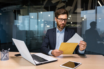Mature and serious businessman inside office thoughtfully reading letter received notification, man in business suit investor at work using laptop.