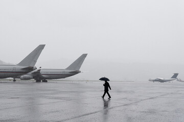 Naklejka premium A man under an umbrella at the airport. Desert airfield. Rainy weather at the airport. Large airfield with aircraft. The man goes to the plane.
