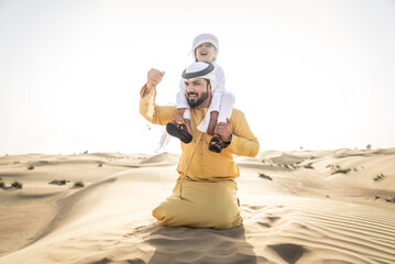 Arabian man and son wearing traditional emirates dishdasha and playing in the desert - Middle eastern family portrait in Dubai desert