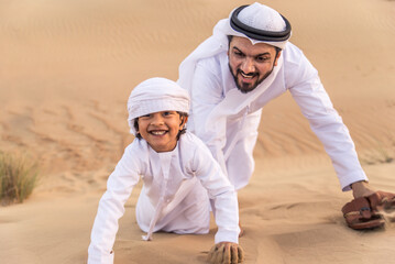Fototapeta na wymiar Playful arab man and his son wering traditional middle eastern emirate clothing playing and having fun in the desert of Dubai