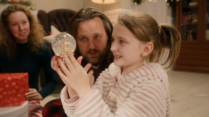 Happy caucasian family opening gifts on Christmas under decorated Christmas tree. Young girl unpacks glass magic snow globe. Warm atmosphere at home on Christmas or New Year. Winter holidays.