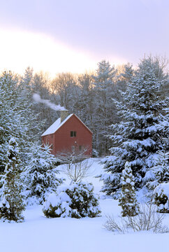 Tranquil New England Winter Scene Following A Departing Snowstorm. Clearing Sky, Warm Cozy Home With Smoke Rising From Stone Chimney, And Trees Covered In Fresh Snow.