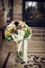 A wedding bouquet in a glass vase yellow white and pink flowers with beige milky ribbons stands on the floor in a room with a brick wall by the fireplace brown background
