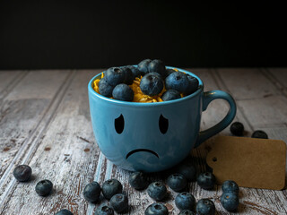 Close-up of a blue mug with a sad face drawn on it filled with blueberries and cereal, blue monday...