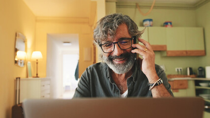 Mature businessman working on laptop, talking on mobile phone sitting in home kitchen