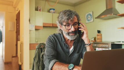 Mature businessman working on laptop, talking on mobile phone sitting in home kitchen