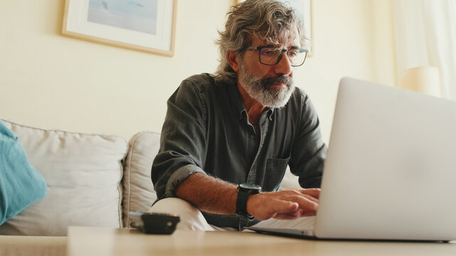 Elderly positive man typing on laptop sitting on sofa in living room