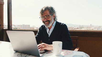 Happy middle aged man communicates remotely using laptop. Working from home office