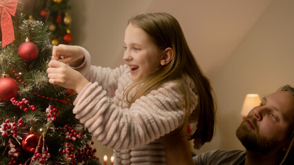 Father lift his young daughter up to put Christmas ball on tree. Happy family decorating Christmas tree and preparing home together for Christmas or New Year. Festive decorations for winter holidays.