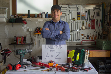 Image of a smiling man in his workshop behind a bench full of work tools with handyman from A to Z written on it. Man who can fix and fix everything
