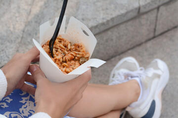 Woman holding paper box of takeaway noodles with fork outdoors, closeup. Street food