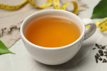 Cup of herbal diet tea and measuring tape on light marble table, closeup. Weight loss concept