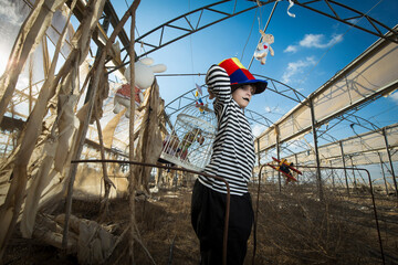 a boy with a sinister smile in a clown costume plays and has fun in an abandoned greenhouse with a parrot in a cage and plush toys