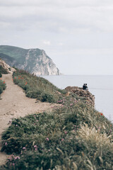 Path By The Sea. A couple sits on a stone wall overlooking the ocean and a large cliff under a cloudy sky. Coastal Cliffside Path With Ocean View Under Bright Daylight
