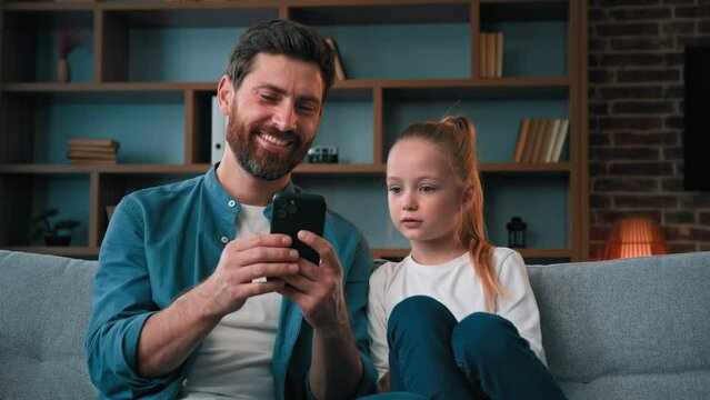 Cute small baby girl child resting on cozy couch with caring young father man playing game on smartphone watching media content on phone using mobile apps shopping online taking family selfie together