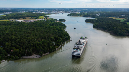 Fototapeta premium Luxury cruise vessel making way ahead in beautiful Finnish archipelago. Narrow fairway and shoreline proximity. High altitude aerial stern view.
