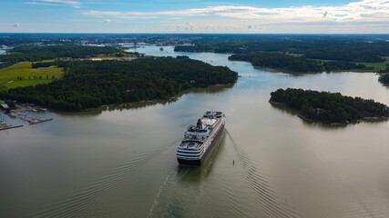Luxury cruise vessel making way ahead in beautiful Finnish archipelago. Narrow fairway and shoreline proximity. High altitude aerial stern view.