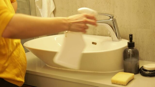 Female hand spray detergent on bathroom sink surface then scrub with sponge. Wiping sink and water tap with cleanser.