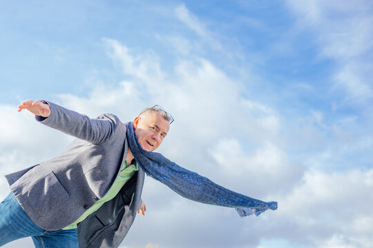 Portrait of handsome middle-aged man balancing on one leg on cloudy sky background, stretching hands, looking at camera.