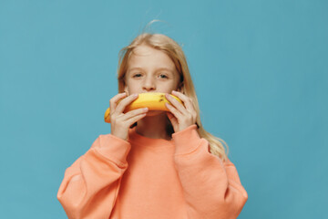  handsome, happy girl stands in orange clothes on a blue background and holds a banana in her hand, substituting it as a smile to her face. Studio photo with empty space for advertising insert