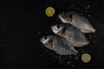 raw gutted fish lies on a stone cutting board with coarse salt and lemon. sea bream on a black background. top view with copy space